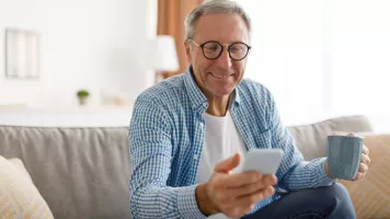 gray haired white man with glasses on couch holding a coffee cup and looking at a phone