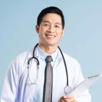A dark-haired doctor wears a labcoat and smiles, standing against a blue background