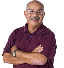 Older hispanic man with glasses and a gray mustache. His arms are crossed in a relaxed manner and he's wearing a short sleeve maroon polo shirt and a watch. 