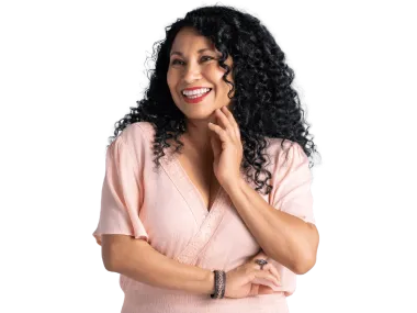 Woman with dark curly hair smiles and touches her face