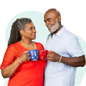 African American couple gazing lovingly at each other and clinking mugs. Woman is wearing a red shirt and holding a blue mug, man is wearing a light blue shirt and holding a red mug.
