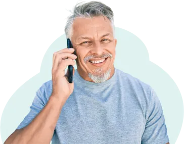 A man with silver hair swept across his forehead is smiling while talking on the phone. He wears a soft blue jersey cotton shirt and looks happy to be having the conversation.