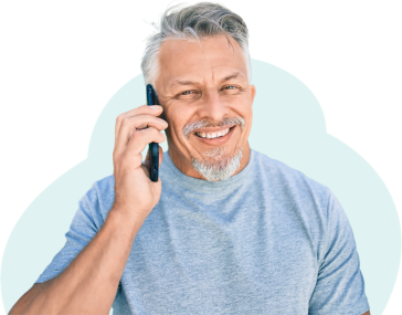 A man with silver hair swept across his forehead is smiling while talking on the phone. He wears a soft blue jersey cotton shirt and looks happy to be having the conversation.