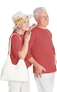 Caucasian couple in their late 60's wearing red shirts and white pants. Woman is behind the man, leaning on his shoulder and carrying a white tote bag over her shoulder. She is wearing a white broad brim straw hat with yellow flowers on the hair band. Both are smiling and looking to the right. 